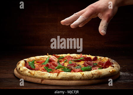 Chef's Hand streuen spicies über köstliche italienische Pizza Margherita mit Tomaten, Basilikum und hamon serviert auf hölzernen Platte über Rustikal backgr Stockfoto