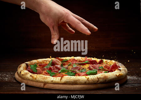 Chef's Hand streuen spicies über köstliche italienische Pizza Margherita mit Tomaten, Basilikum und hamon serviert auf hölzernen Platte über Rustikal backgr Stockfoto