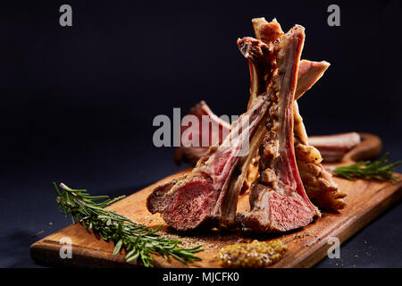Lammrücken mit Rosmarin, mit einigen spicies auf Holz Schneidebrett über dunklen Hintergrund verschüttet, Seitenansicht, selektiver Fokus, geringe Tiefenschärfe Stockfoto