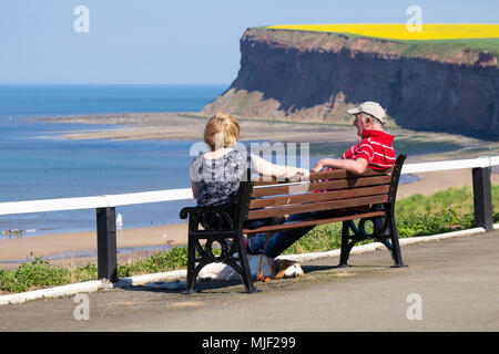 Rückansicht eines Paares mit Blick auf das Meer und einem Hund im Schatten unter der Bank. VEREINIGTES KÖNIGREICH Stockfoto