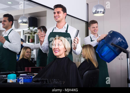 Lächelnd dunkelhaarigen Mann tun Frisur für Happy reife blonde Frau im Friseursalon Stockfoto