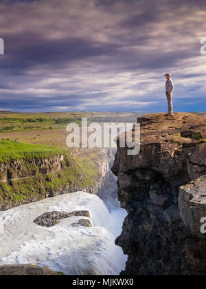 Junge Menschen stehen am Rand einer Klippe mit Blick auf den Horizont, über einen Wasserfall gegen eine dramatische Wolkenhimmel, Gullfoss, Island Stockfoto