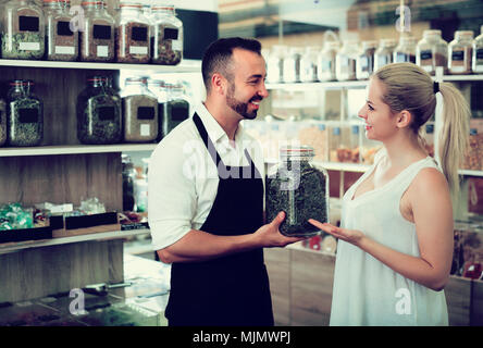 Freundlich lächelnden Mann mit Schürze Verkauf von Kräutern und Gewürzen zu einer weiblichen Kunden im Bioladen Stockfoto