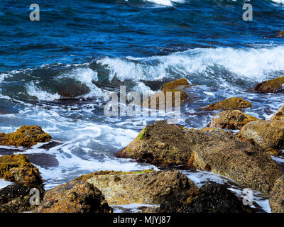 Spritzer aus den Wellen stoßen gegen die felsige Küste, das Meer ist blau Stockfoto