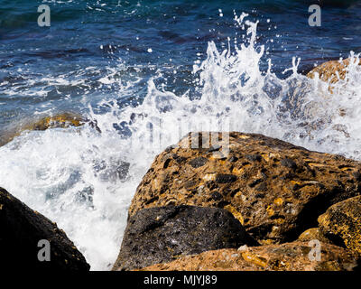 Spritzer aus den Wellen stoßen gegen die felsige Küste, das Meer ist blau Stockfoto