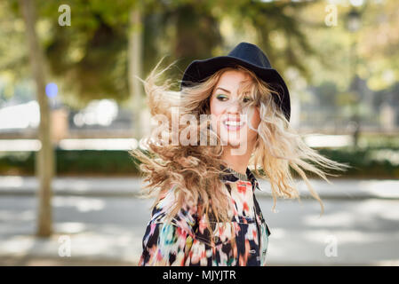 Junge Frau mit fliegendem Haar zu tragen Jacke und Hut, im städtischen Hintergrund glücklich Stockfoto