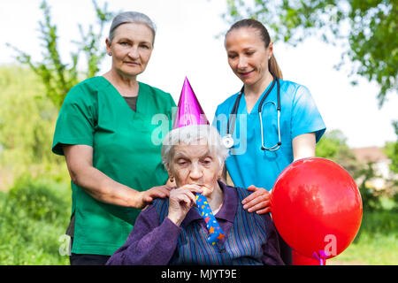 Ältere behinderte Frau in einem Rollstuhl feiert Geburtstag mit freundlichen weiblichen Pflegepersonen im Freien Stockfoto