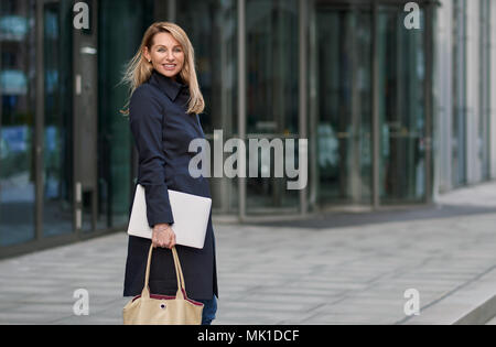 Attraktive stilvolle Blondine Geschäftsfrau zu Fuß durch die Stadt anhalten zurück in die Kamera mit einer warmen, freundlichen Lächeln vor einem Bürogebäude zu suchen Stockfoto