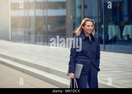 Attraktive stilvolle blonde Frau verlassen Ihren Arbeitsplatz als sie Spaziergänge eine ruhige high key Urban Street lächelnd, als sie ihren Laptop trägt mit Kopie Raum Stockfoto