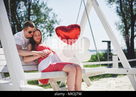 Warten Baby. schwangere Frau mit dem geliebten Mann sitzen auf einer Bank. Mann Hand umfasst einen runden Bauch Frau. Elternschaft. schwangere Frau in einem weißen Kleid in Kirsche mit einem roten Bogen. close-up. Neun Monate Stockfoto