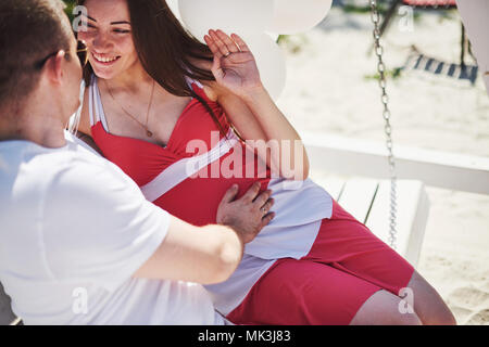 Warten Baby. schwangere Frau mit dem geliebten Mann sitzen auf einer Bank. Mann Hand umfasst einen runden Bauch Frau. Elternschaft. schwangere Frau in einem weißen Kleid in Kirsche mit einem roten Bogen. close-up. Neun Monate Stockfoto