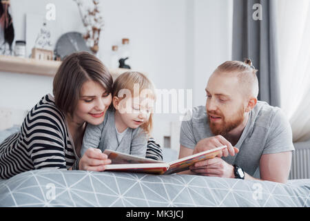 Vater, Mutter und Tochter lesen Kinderbuch auf einem Sofa im Wohnzimmer. Gerne große Familie lesen Sie ein interessantes Buch über einen festlichen Tag. Die Eltern lieben ihre Kinder Stockfoto