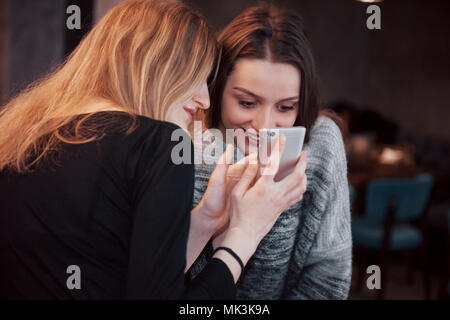 Zwei lächelnde Freunde lesen lustige online auf modernen Telefon sitzend mit leckeren Kaffee im Restaurant chatten. Hipster Mädchen genießen Freizeit im Cafe mit heißen Getränken und Messaging auf zelluläre Stockfoto