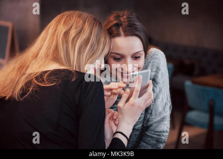 Zwei lächelnde Freunde lesen lustige online auf modernen Telefon sitzend mit leckeren Kaffee im Restaurant chatten. Hipster Mädchen genießen Freizeit im Cafe mit heißen Getränken und Messaging auf zelluläre Stockfoto