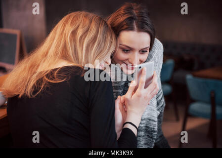 Zwei lächelnde Freunde lesen lustige online auf modernen Telefon sitzend mit leckeren Kaffee im Restaurant chatten. Hipster Mädchen genießen Freizeit im Cafe mit heißen Getränken und Messaging auf zelluläre Stockfoto