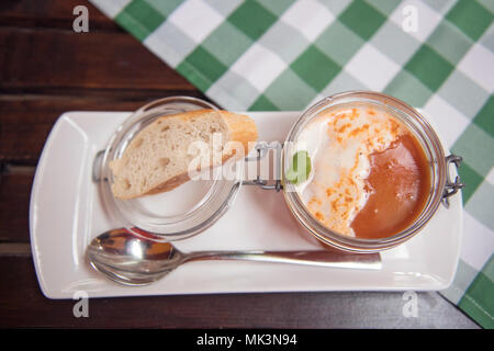 Eine köstliche Portion cremiger Tomatensuppe in einem Glas, garniert mit Basilikum, begleitet von einem Stück frischem Brot auf einem weißen Teller, Stockfoto