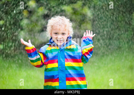 Kleine blonde lockige Junge in eine wasserdichte Jacke in Rainbow Color spielen im Regen. Kinder Spaß im Freien im Herbst. Stockfoto