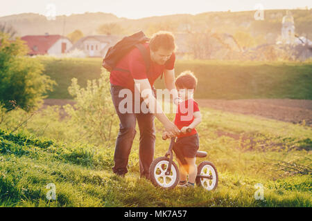 Vater lehrt, sein Sohn, ein Gleichgewicht mit dem Fahrrad in die Landschaft bei Sonnenuntergang zu reiten. Führen Sie Fahrrad ohne Pedale Stockfoto