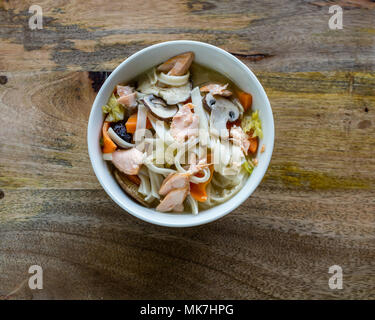 Asiatische Suppe Reis Nudeln Lachs und Gemüse in Weiß Schüssel gegen Holz- Hintergrund Stockfoto