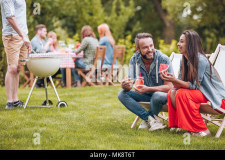 Lächelnde Frau essen Wassermelone und ihr Freund trinken einen Cocktail im Garten während einer Partei Stockfoto