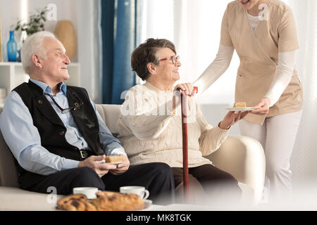 Geriatrische Paar mit Arthritis sitzen auf einer Couch und serviert ein Stück Kuchen während der Wartezeit für Termin beim Arzt in einem luxuriösen privaten clini Stockfoto