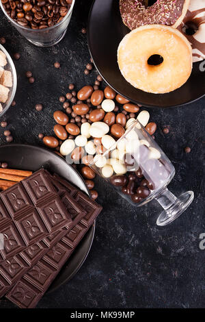 Blick von oben auf die Erdnüsse in Schokolade, auf der dunklen Kammer verschüttet, neben Schokolade Tabletten, Donuts, brauner Zucker und Kaffee Bohnen Stockfoto