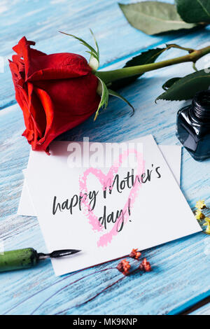 Nahaufnahme einer roten Rose, ein Tintenfass, eine nib Stift und ein Stück Papier mit dem Text zufrieden Muttertag in Es geschrieben, auf blauem rustikalen Holztisch Stockfoto