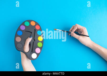Weibliche Hände halten Kunst Palette mit Farben und Pinsel gegen den blauen Hintergrund minimal kreatives Konzept. Stockfoto