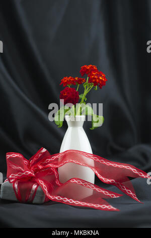 Besondere Geschenk mit roter Schleife mit Rot lantana Blumen in Weiß Vase gegen Schwarz Satin Hintergrund gewickelt hat freudige Stimmung von Romantik und celeb Stockfoto