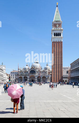 Frau mit rosa Schirm vor St Marks Dom und Campanile in Piazza San Marco, Venedig, Venetien, Italien mit Touristen im Hintergrund Stockfoto