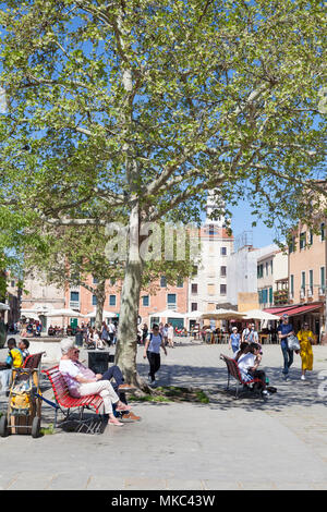 Einheimische und Touristen entspannen auf Bänken unter einem schattigen Baum im Frühling in Campo Santa Margherita, Dorsoduro Venedig, Italien in der Sonne Stockfoto