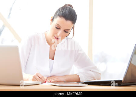 Portrait von lächelnden jungen Verkäuferin Frau tun etwas Schreibarbeit während im Büro Schreibtisch vor dem Laptop sitzen und online arbeiten. Home Office. Stockfoto