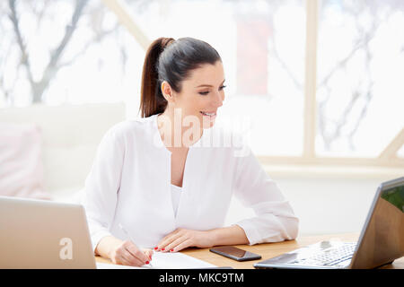 Lächelnd vertrieb Frau dabei einige Unterlagen im Büro sitzend vor Laptop im Büro. Stockfoto