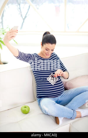 Gerne schwangere Frau die Hand ihr 3D-Ultraschallbild und unter selfie während auf dem Sofa zu Hause sitzen. Stockfoto