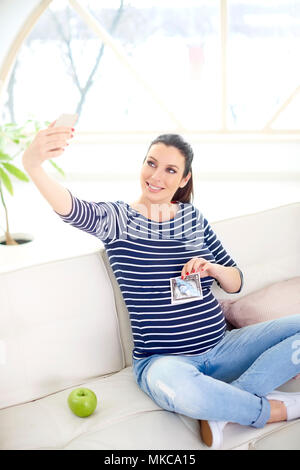 Gerne schwangere Frau die Hand ihr 3D-Ultraschallbild und unter selfie während auf dem Sofa zu Hause sitzen. Stockfoto