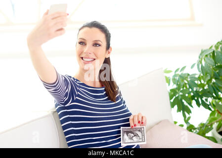 Gerne schwangere Frau die Hand ihr 3D-Ultraschallbild und unter selfie während auf dem Sofa zu Hause sitzen. Stockfoto