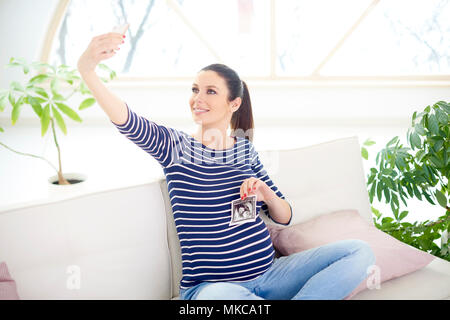 Gerne schwangere Frau die Hand ihr 3D-Ultraschallbild und unter selfie während auf dem Sofa zu Hause sitzen. Stockfoto