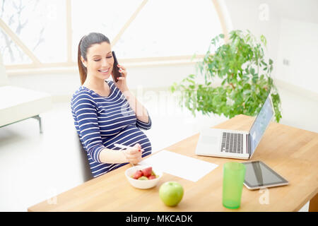 Glückliche junge schwangere Frau im Gespräch mit jemand auf Ihr Handy und schreiben etwas beim Sitzen am Schreibtisch vor dem Laptop. Stockfoto