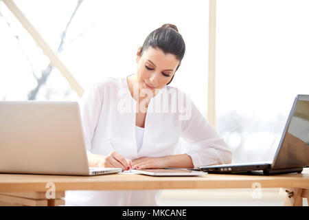 Schöne junge Frau im Büro Schreibtisch hinter Computern sitzen und und tun etwas Schreibarbeit, während Ihr Unternehmen verwalten. Home Office. Stockfoto