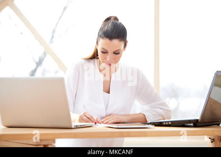 Schöne junge Frau im Büro Schreibtisch hinter Computern sitzen und und tun etwas Schreibarbeit, während Ihr Unternehmen verwalten. Home Office. Stockfoto