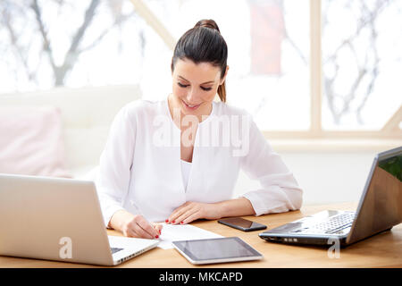 Schöne junge Frau im Büro Schreibtisch hinter Computern sitzen und und tun etwas Schreibarbeit, während Ihr Unternehmen verwalten. Home Office. Stockfoto