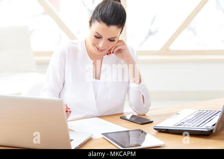 Schöne junge Frau im Büro Schreibtisch hinter Computern sitzen und und tun etwas Schreibarbeit, während Ihr Unternehmen verwalten. Home Office. Stockfoto
