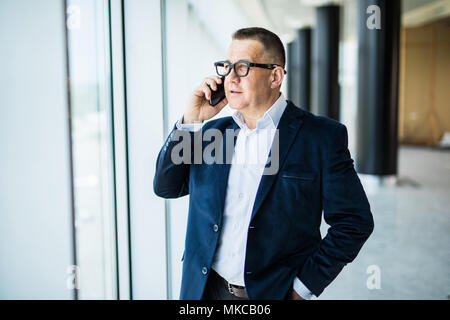 Lächelnd im mittleren Alter Geschäftsmann mit Mobiltelefon sitzen vor dem Fenster mit Blick auf die Stadt Stockfoto