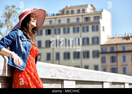 Lächelnd touristische Frau in hat Schiefe auf Handlauf und weg schauen auf der sonnigen Stadt Straße. Stockfoto
