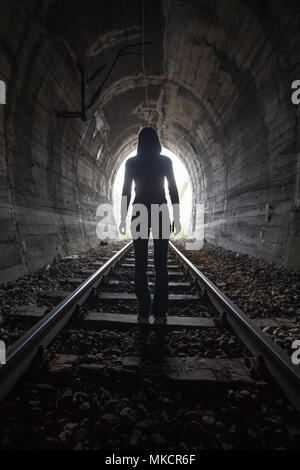 Mann Silhouette, die in einem Tunnel in der Mitte der Gleise mit Blick auf das Licht am Ende des Tunnels in einem konzeptionellen Bild Stockfoto