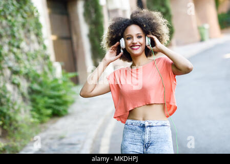 Porträt der jungen attraktiven schwarzen Mädchen im städtischen Hintergrund die Musik mit Kopfhörern. Frau orange T-Shirt und eine blaue Jeans mit einem Stockfoto