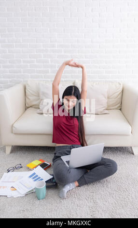 Asiatische Frau arm bis zu sich selbst Stretch nach Laptop Computer im Wohnzimmer zu Hause zu arbeiten. die Frau wieder am Sofa sitzen auf dem Teppich an der Wand arbeiten Stockfoto