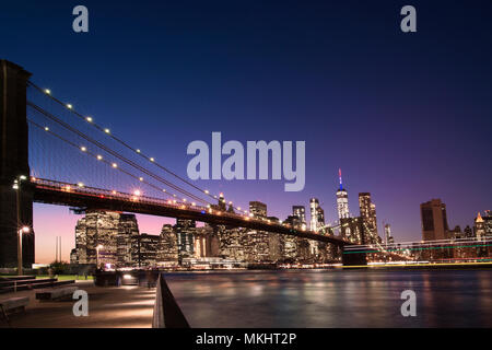 Brooklyn Bridge und die beleuchtete Skyline von Manhattan am Abend mit blauem Himmel und glatte Wasseroberfläche geschossen aus Brooklyn, New York, USA. Stockfoto