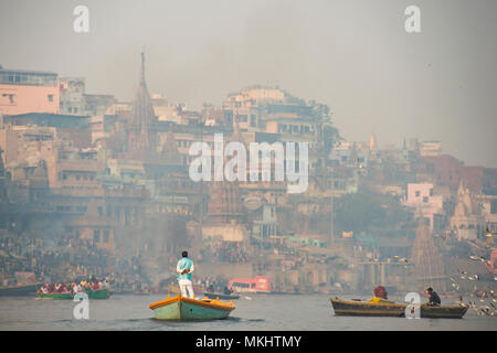 Ein Mann ist Segeln auf sein hölzernes Boot in den heiligen Fluss Ganges in der Stadt Varanasi bei Sonnenuntergang. Verschwommen Manikarnika Ghat für den Hintergrund. Stockfoto