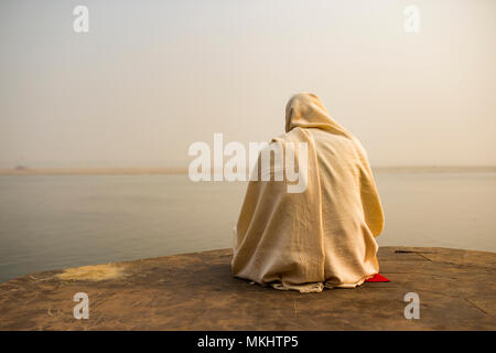 Ein heiliger Mann in Weiß gekleidet ist betend und meditierend auf einem der vielen Ghats von Varanasi vor dem heiligen Fluss Ganges in Indien. Stockfoto